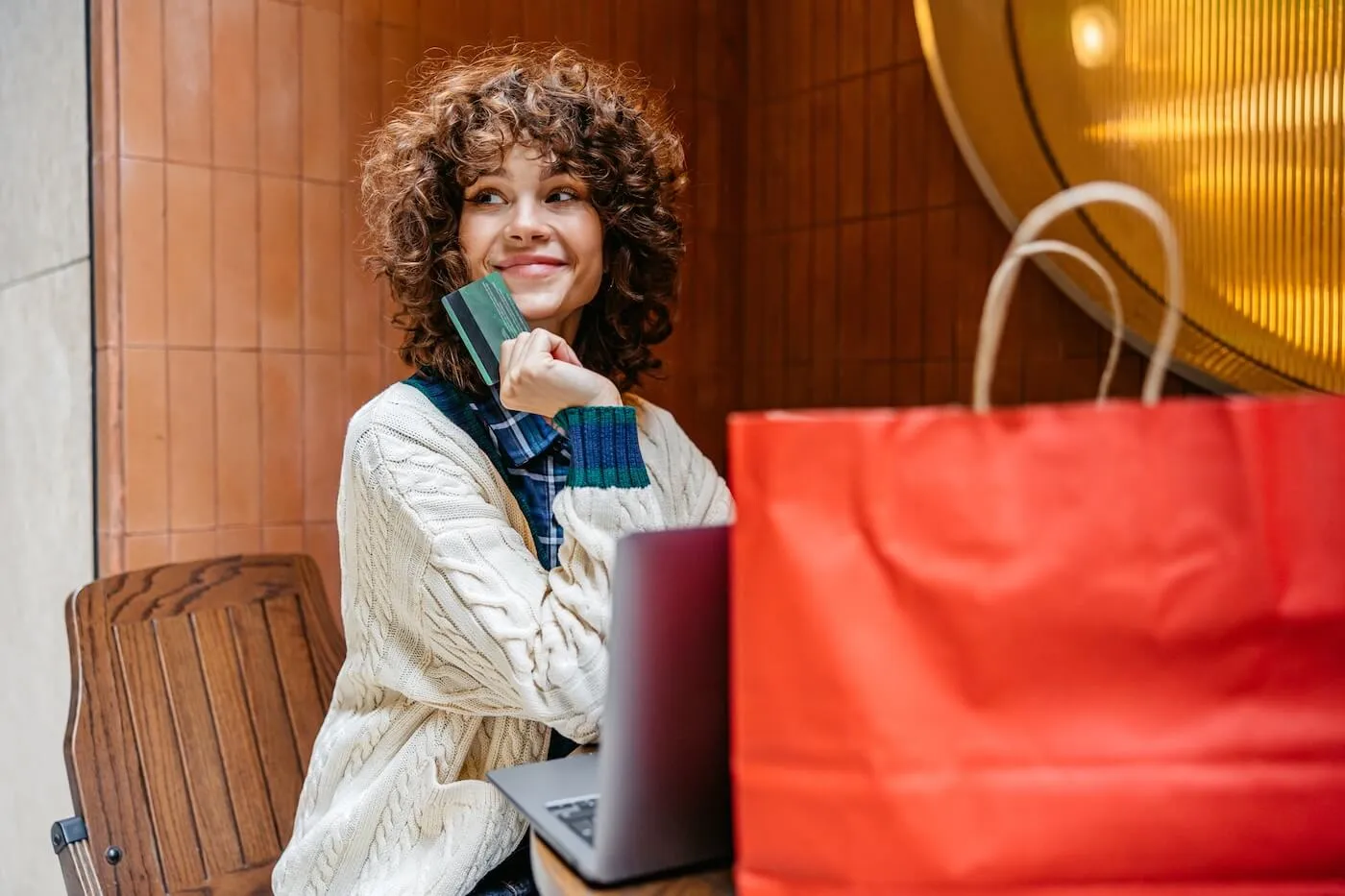 Cheerful woman holding a credit card and browsing online deals on her laptop with a red shopping bag by her side.