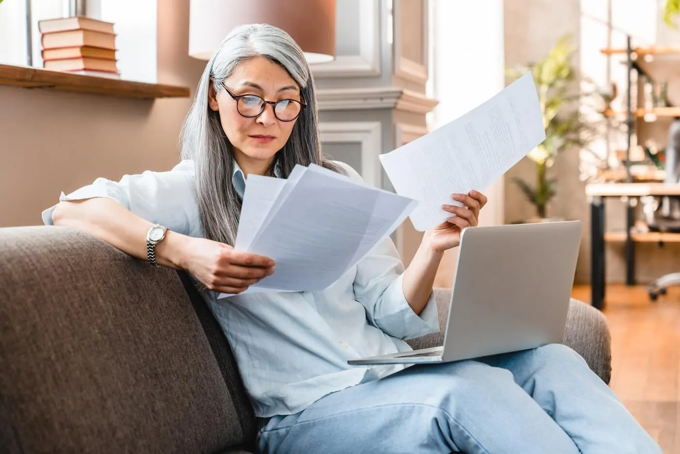 Focused mature woman reviewing the printouts while sitting on a couch with a laptop